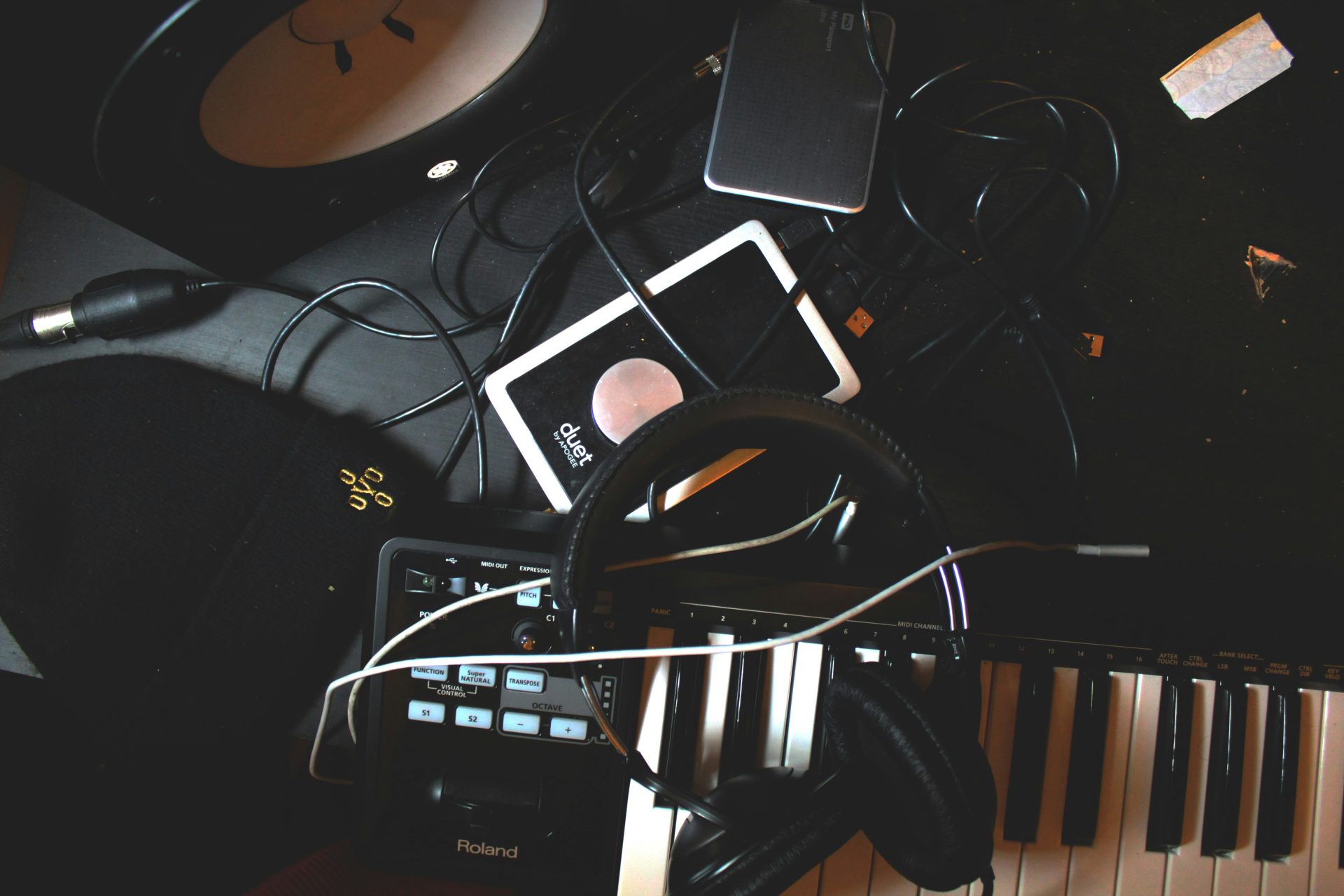 An overhead shot of a cluttered desk features a Roland keyboard, headphones, an Apogee Duet interface, and various cables.