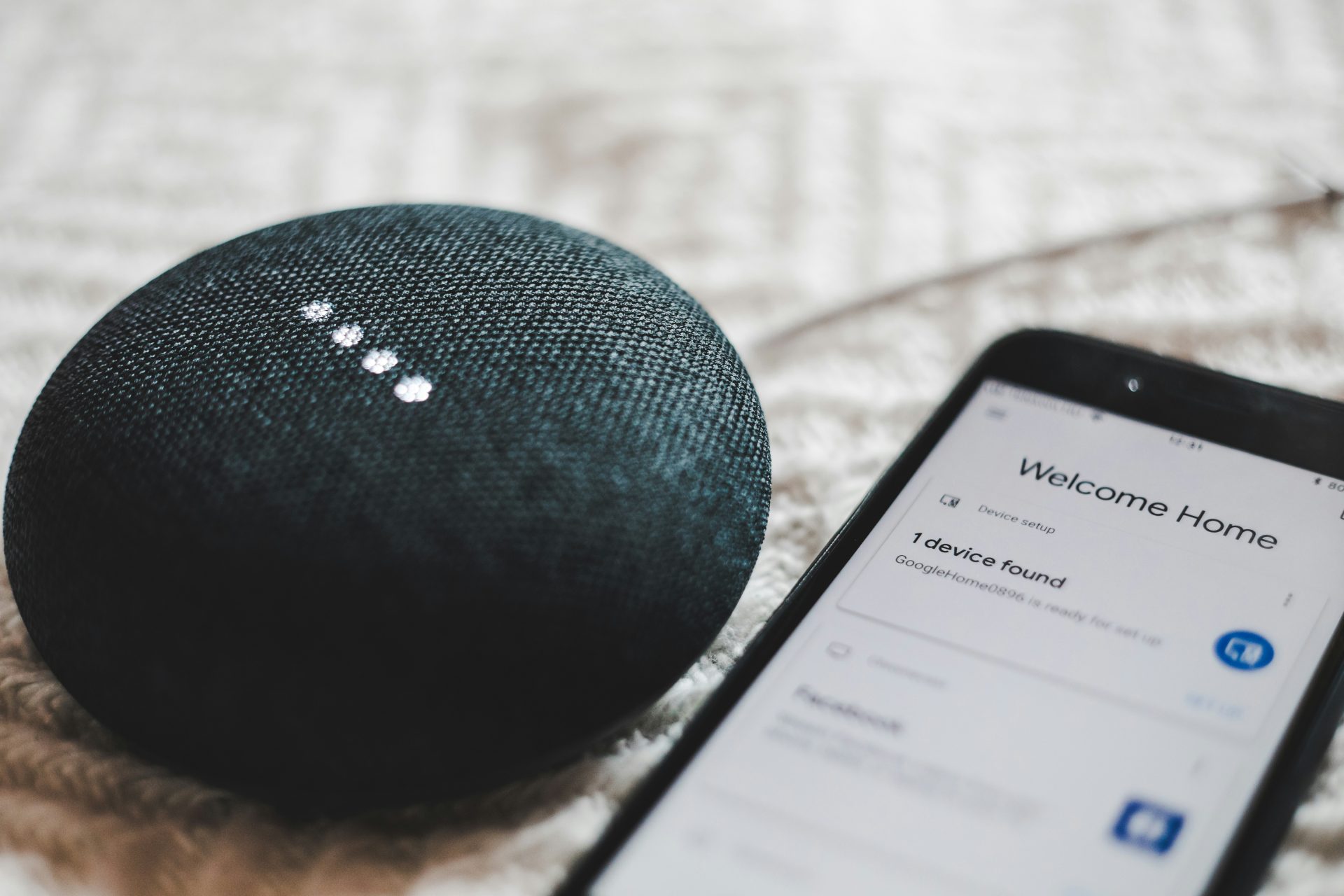 A black Google Nest Mini smart speaker with illuminated dots sits beside a smartphone displaying the "Welcome Home" setup screen for a new device.