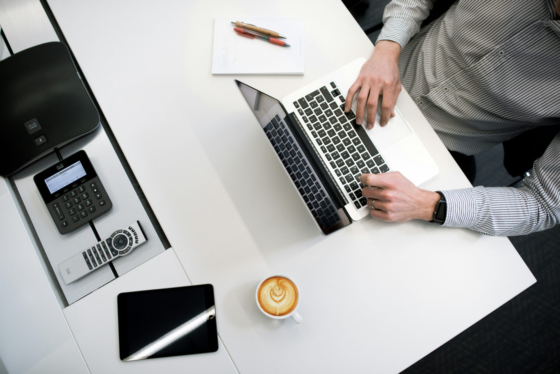 An overhead shot of a person working on a laptop at a white desk, with a coffee cup, tablet, and various office electronics nearby.