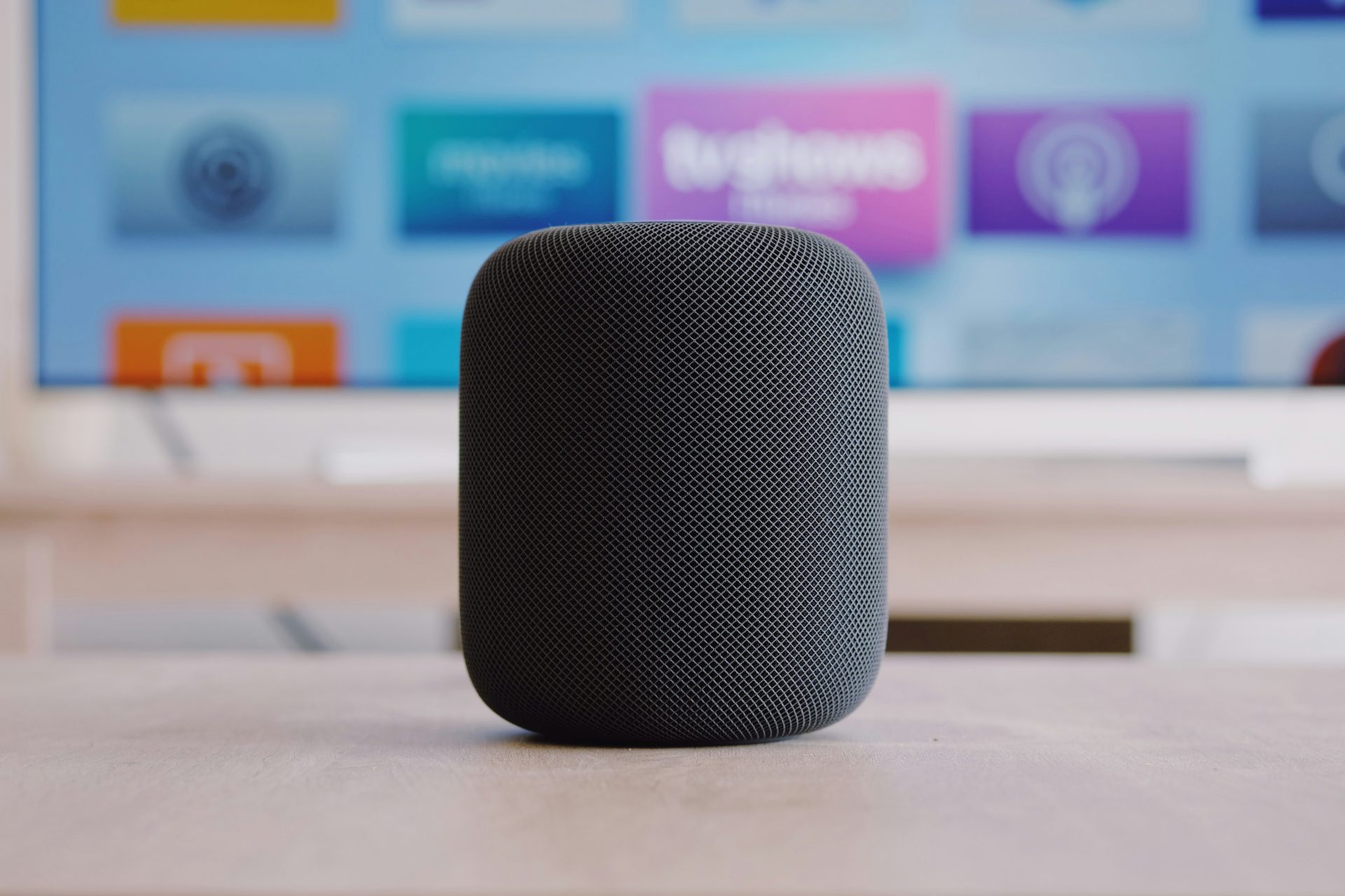 A black Apple HomePod smart speaker sits on a wooden surface in front of a blurred television screen displaying app icons.