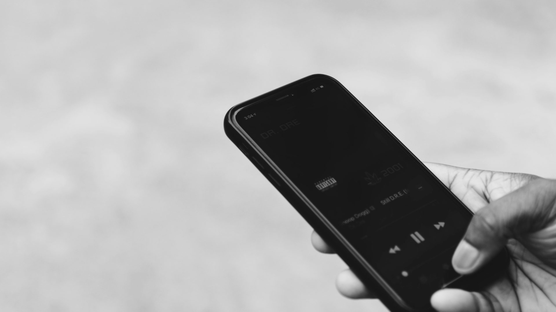 A hand holds an iPhone displaying a music player interface for "Still D.R.E." by Dr. Dre, in black and white.